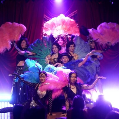 Group of dancers on a stage with large colorful feather fans (pink, blue, green) under purple lights; audience silhouettes in foreground.