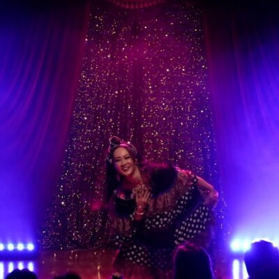 Performer on a glittery stage, bending forward with a smile, wearing polka-dot attire and a headwrap, purple stage lights in the background.