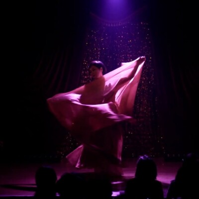Dancer in a pink flowing skirt twirls on stage under a purple spotlight, with silhouetted audience in the foreground.