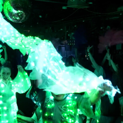 Crowd at a nightclub dancing in bright green LED costumes with illuminated fabric swirling above, disco ball overhead.