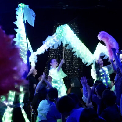 Stage performance featuring dancers in glowing LED costumes waving a lit dragon prop amid a cheering crowd in a dark theater, with blue and green lights