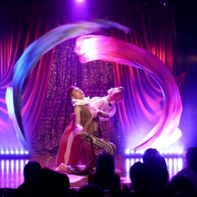 A performer in a colorful costume kneels on stage, twirling long rainbow ribbons in a circular arc under purple stage lights, with a glittery backdrop and audience silhouettes watching.