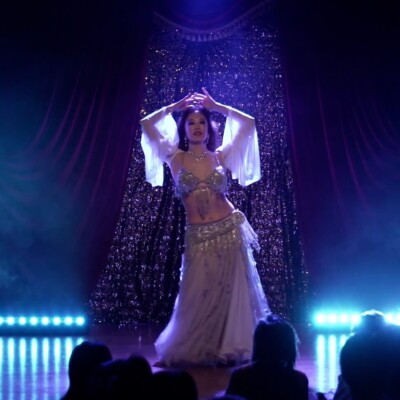 Belly dancer in a sparkling silver outfit performs on a lit stage with blue lights and velvet curtains, audience in foreground.