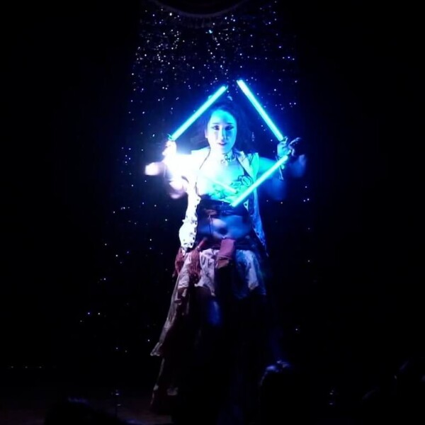 Performer on a dark stage holding two glowing blue light swords crossed in front, against a starry backdrop.