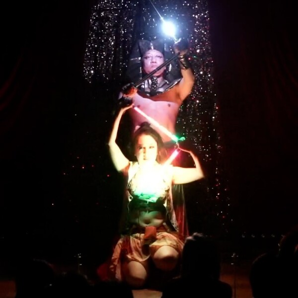 Two performers on a dark stage; a woman kneels in front with glowing neon sticks, a man behind holds a sword and a bright light, glittering backdrop.