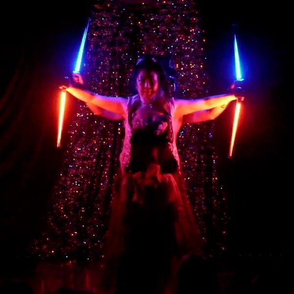 Performer on a dark stage holding red and blue glowing swords, arms outstretched with a glittery backdrop behind.
