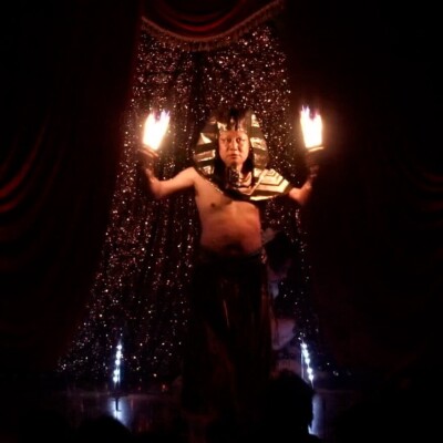 Male stage performer wearing an Egyptian pharaoh headdress and bare chest, holding flaming torches with a glittering backdrop and red curtains behind him.