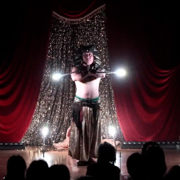 Male performer in a metallic costume and headpiece stands center stage, holding glowing rods as audience watches from the dark foreground.