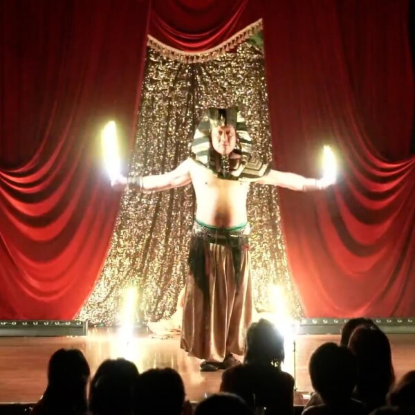 Performer in a pharaoh costume on stage with outstretched arms and glowing torches, red curtains behind him.