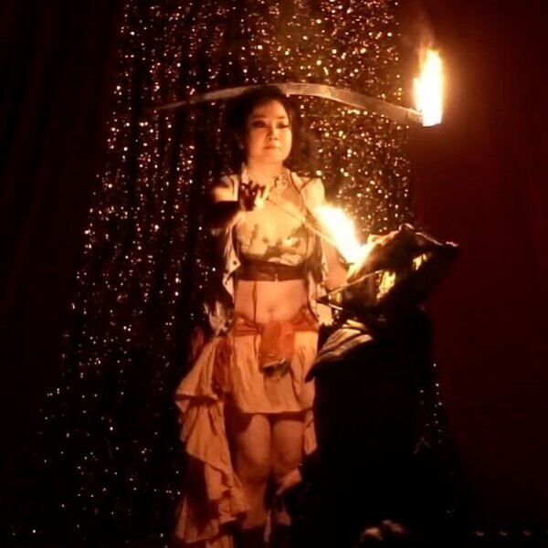 Female fire performer on stage in a pink, ruffled outfit, spinning flaming torches with a sparkly curtain backdrop.