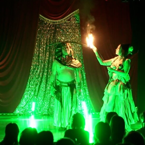 Stage performance with two dancers in Egyptian-inspired costumes; a fire-breathing act under green lighting and a red curtain backdrop.