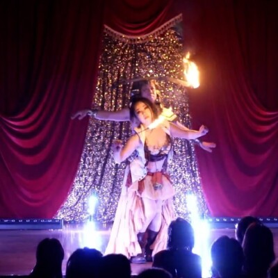 Stage performance with two dancers in flowing costumes in front of a sparkling backdrop; a performer holds a flame, while red velvet curtains frame the scene and the audience watches from the darkened foreground.