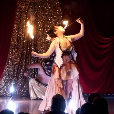 Fire performer in a layered, tattered costume spins flaming torches on a glittery stage backdrop while an audience watches the show.