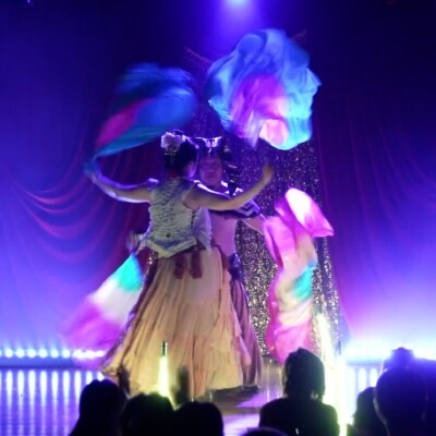 Dancers on stage in flowing dresses twirl colorful scarves under purple stage lights, with audience silhouettes in foreground.