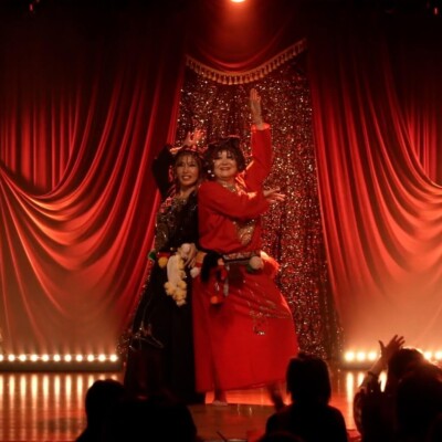 Two performers dance on a glittery red stage with red curtains, wearing festive outfits as the audience watches.