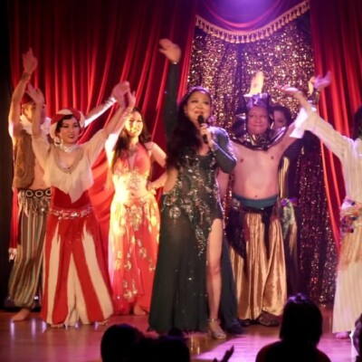 Stage performance with a singer center stage and dancers in colorful, sequined costumes raising their arms under red curtains.
