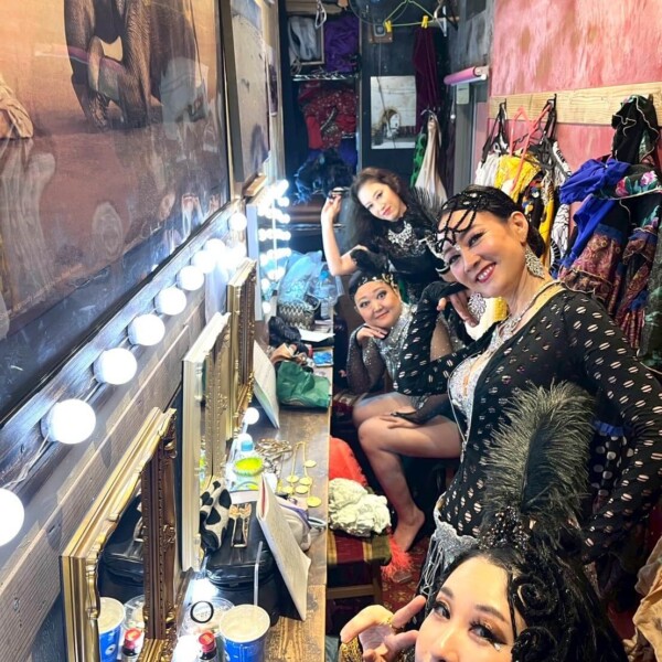 Three women in glittery stage costumes pose in a cluttered backstage dressing room with mirrors and bright bulb lights.