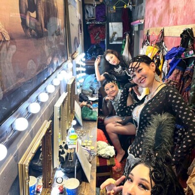 Three women in glittery stage costumes pose in a cluttered backstage dressing room with mirrors and bright bulb lights.